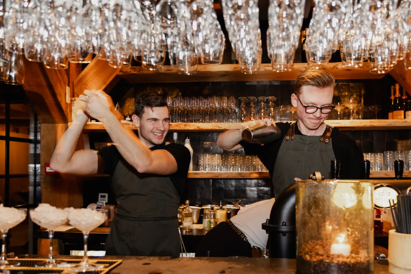 Twee barmannen in zwart met een schort shaken tegelijk achter de bar, onder rijen ondersteboven hangende glazen; op de bar staan drie coupes met schuim en een koperkleurige emmer.