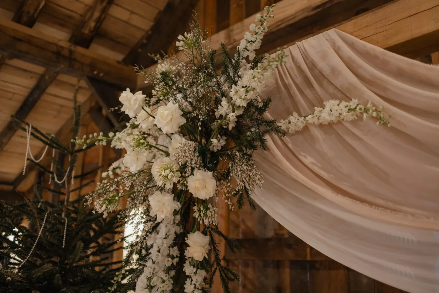 Een close-up van het bloemstuk naast de ceremonieboog in de Barn: witte rozen, witte delphinium en gipskruid tussen donkergroene conifeertakken, tegen de plooiende crème sluierstof die van de houten spanten valt.