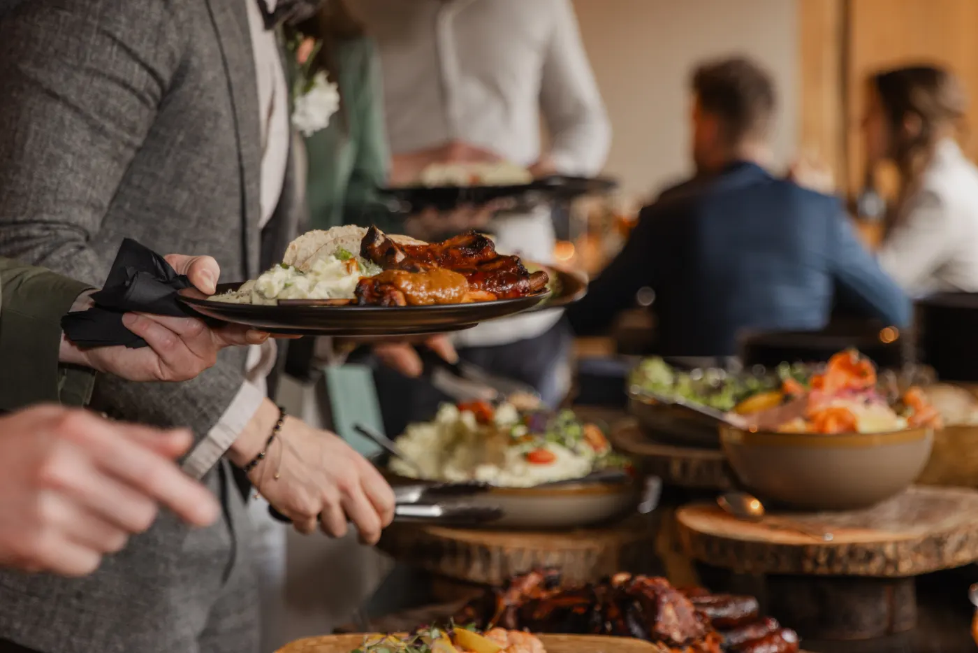 Een gast schept bij het buffet: een bord met gegrild vlees, rijst en salade in de hand, en op de voorgrond houten planken en aardewerken schalen met satéspiesjes, gegrild vlees en salades op boomschijven.