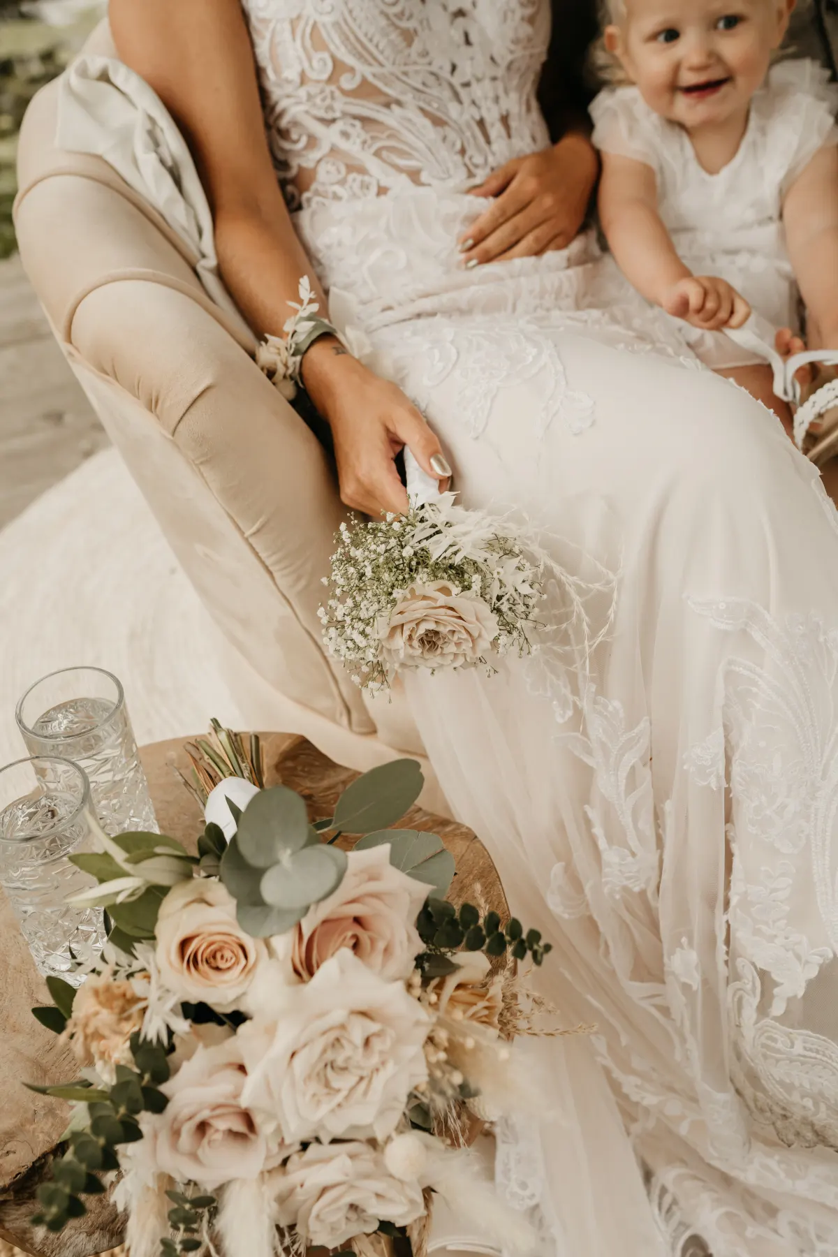 Een bruid zit in een crème fluwelen fauteuil, in beeld vanaf haar schouders: een kanten bovenstuk met bloemapplicatie, een corsage van droogbloemen om haar pols en een klein boeketje blush rozen in haar handen. Rechts naast haar een lachend meisje in een wit kanten jurkje met bungelende benen.