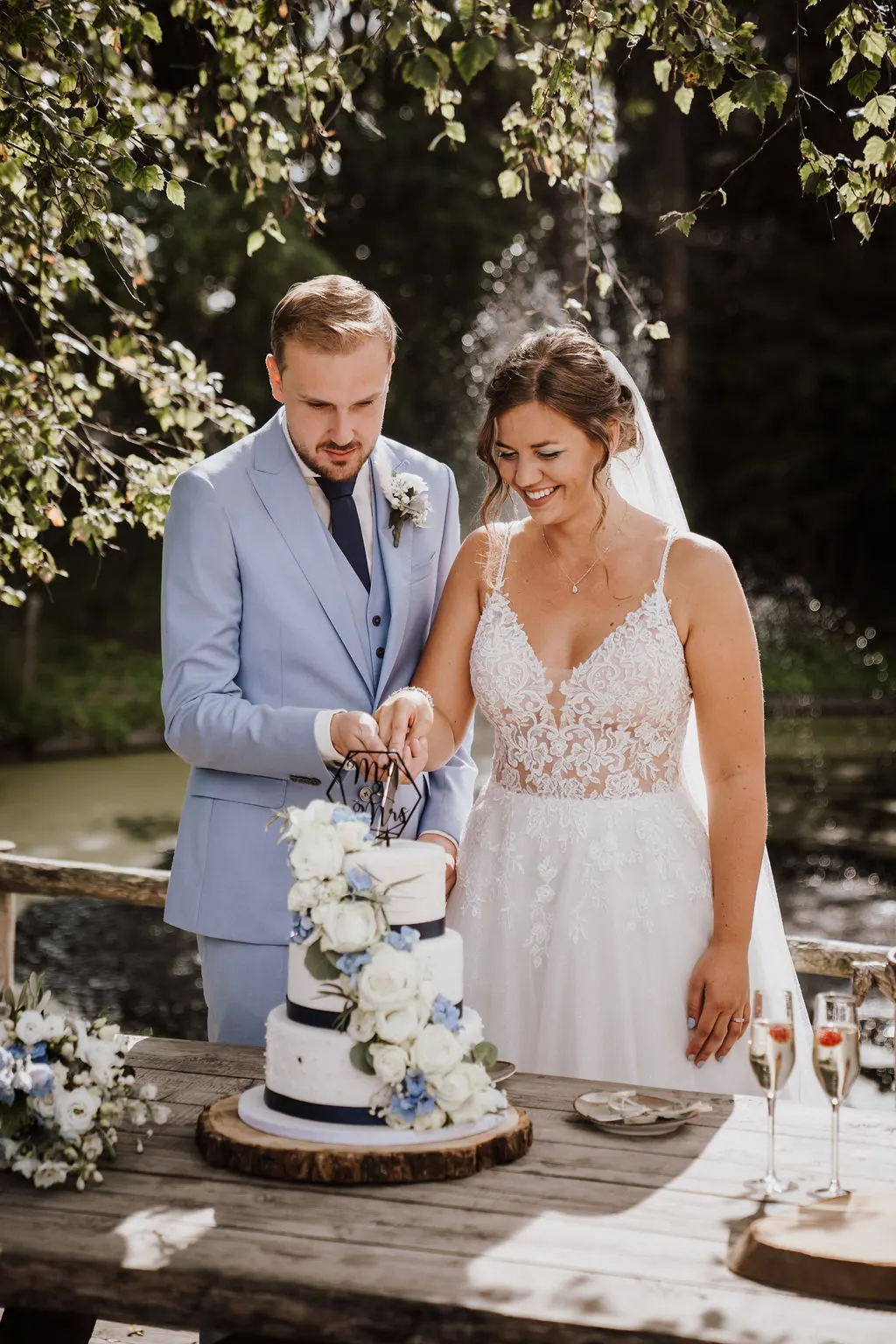 Het bruidspaar snijdt samen de bruidstaart aan bij het water: hij in een lichtblauw driedelig pak, zij in een kanten jurk met sluier, boven een drielaagse witte taart met donkerblauw lint, witte rozen en blauwe hortensia op een houten boomschijf.