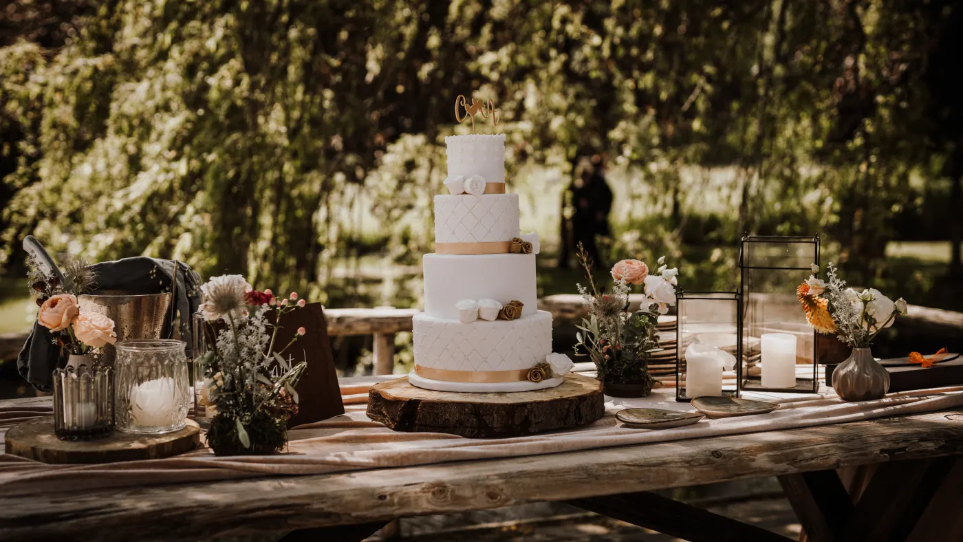 Een drielaagse witte bruidstaart met gouden lint en een gouden topper staat op een houten boomschijf op een verweerde tafel buiten, tussen zwarte lantaarns met stompkaarsen en witte vaasjes met droog- en zomerbloemen.