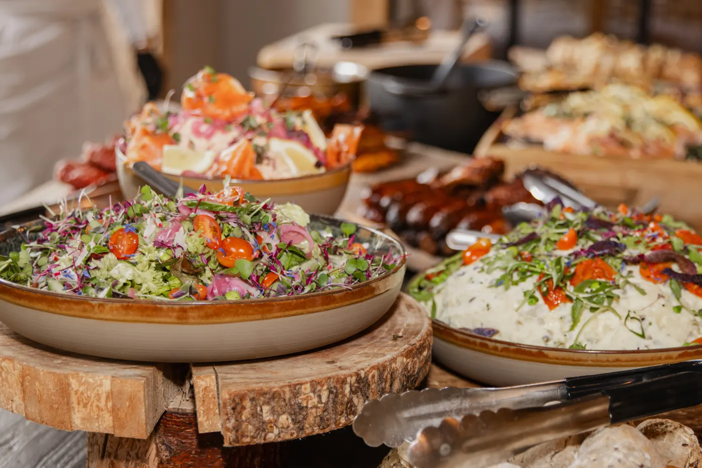 Het buffet van dichtbij: aardewerken schalen met een frisse salade van tomaat en rode ui, rijst en gegrild vlees, geserveerd op houten boomschijven met serveertangen ernaast.