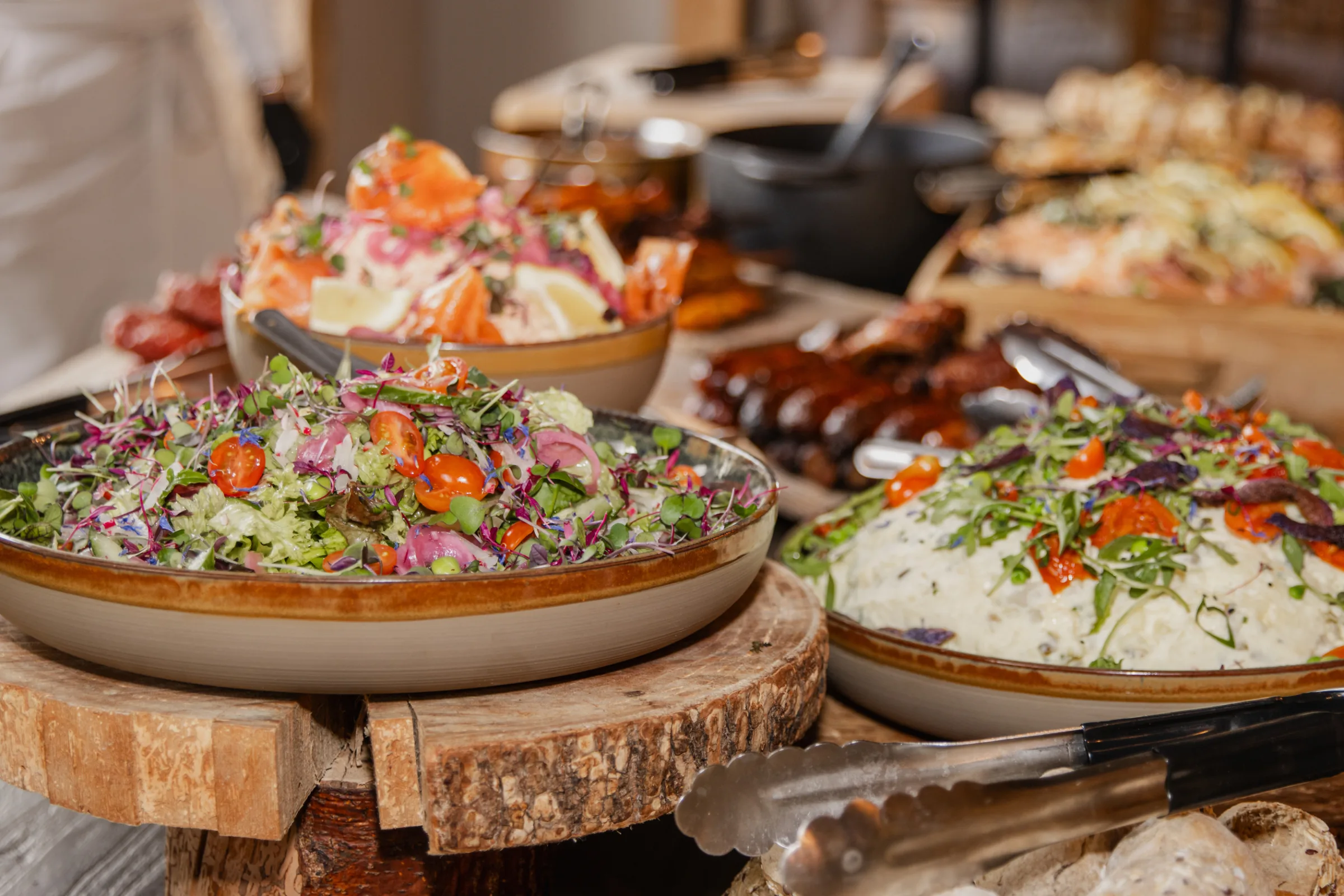 Het buffet van dichtbij: aardewerken schalen met een frisse salade van tomaat en rode ui, rijst en gegrild vlees, geserveerd op houten boomschijven met serveertangen ernaast.