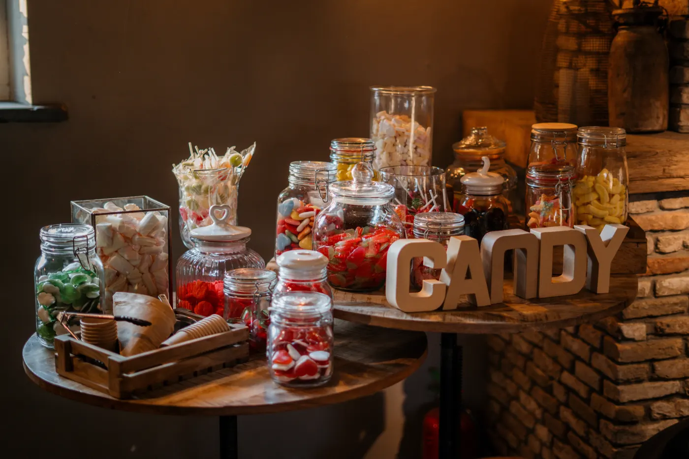 De candy table op twee ronde houten tafels: glazen potten vol snoep, spekjes en zuurtjes, papieren zakjes in een houten kistje en houten letters die CANDY spellen, tegen een donkere wand met een stapel houtblokken.