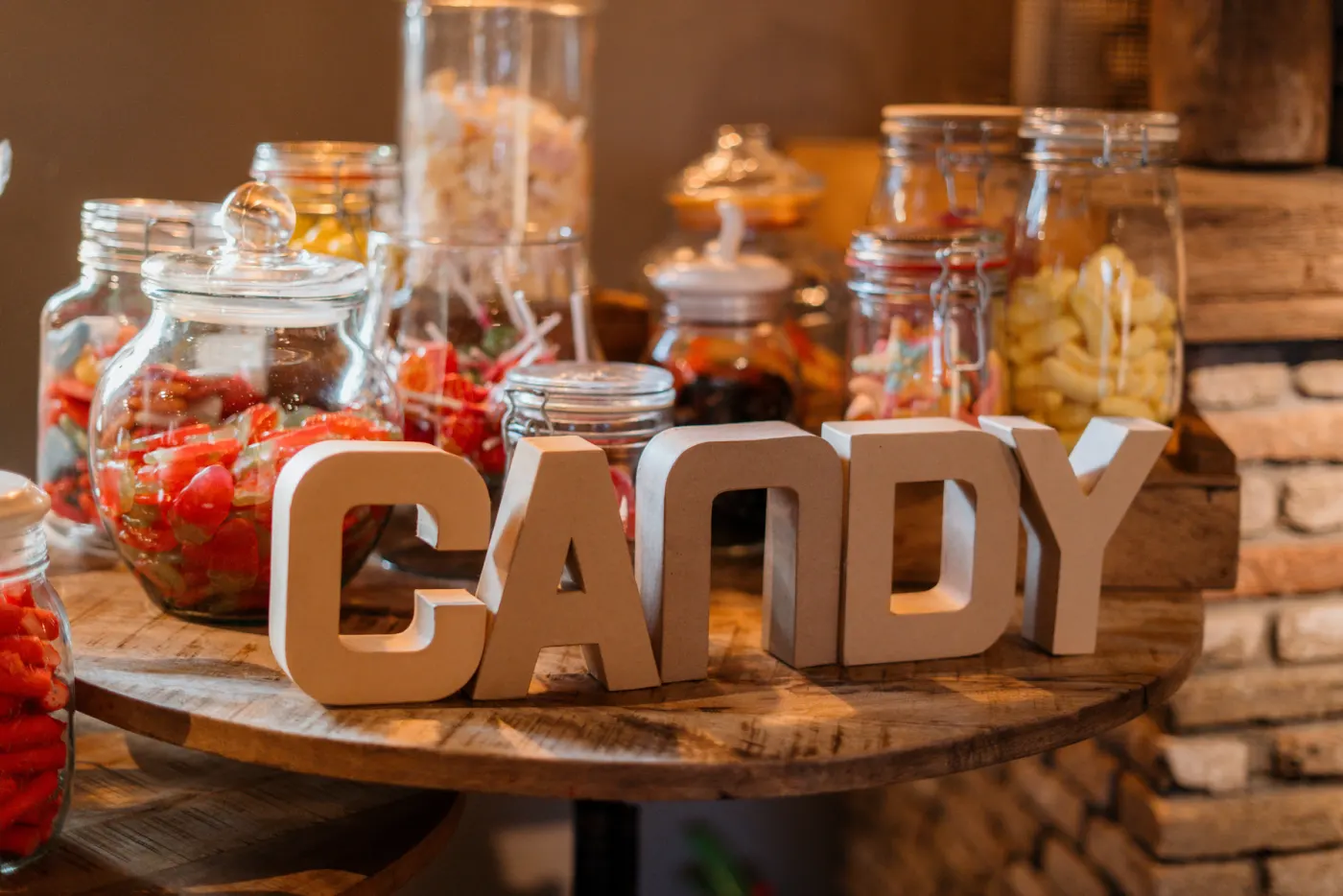 Een candy table: grote glazen stopflessen vol snoep, spekjes en zuurtjes op een houten boomschijf, met houten letters die CANDY spellen ervoor, tegen een donkere houten achtergrond.