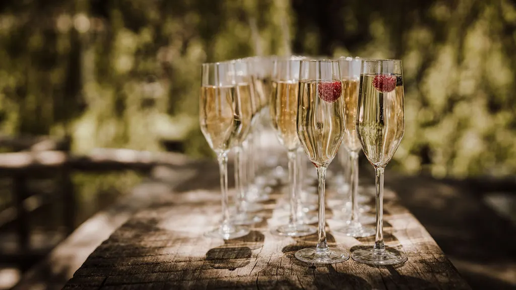 Een rij champagneflûtes met een frambozenbes in elk glas staat in tegenlicht op een verweerde houten tafel buiten, met gouden bokeh van het zomergroen erachter.