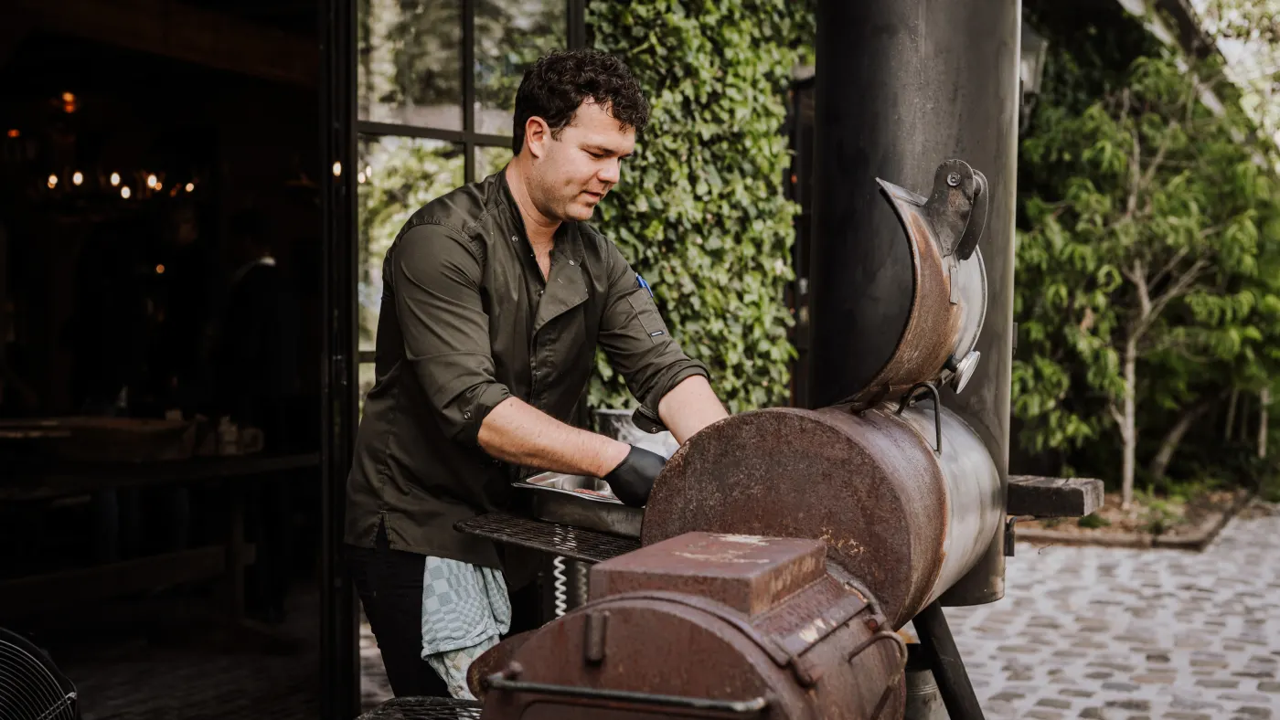 Een chef in een olijfgroen overhemd met een theedoek over zijn arm opent het deksel van een tonvormige smoker, voor een muur begroeid met klimop en zwarte stalen taatsramen.