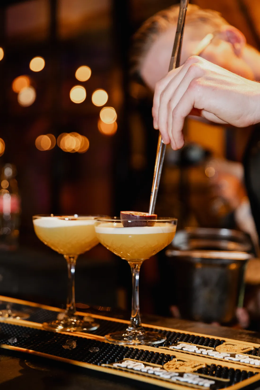 Twee coupecocktails met een schuimlaagje staan op de bar terwijl een barman er met een pincet een garnituur op legt; achter hem de warme bokeh van de lampjes boven de bar.