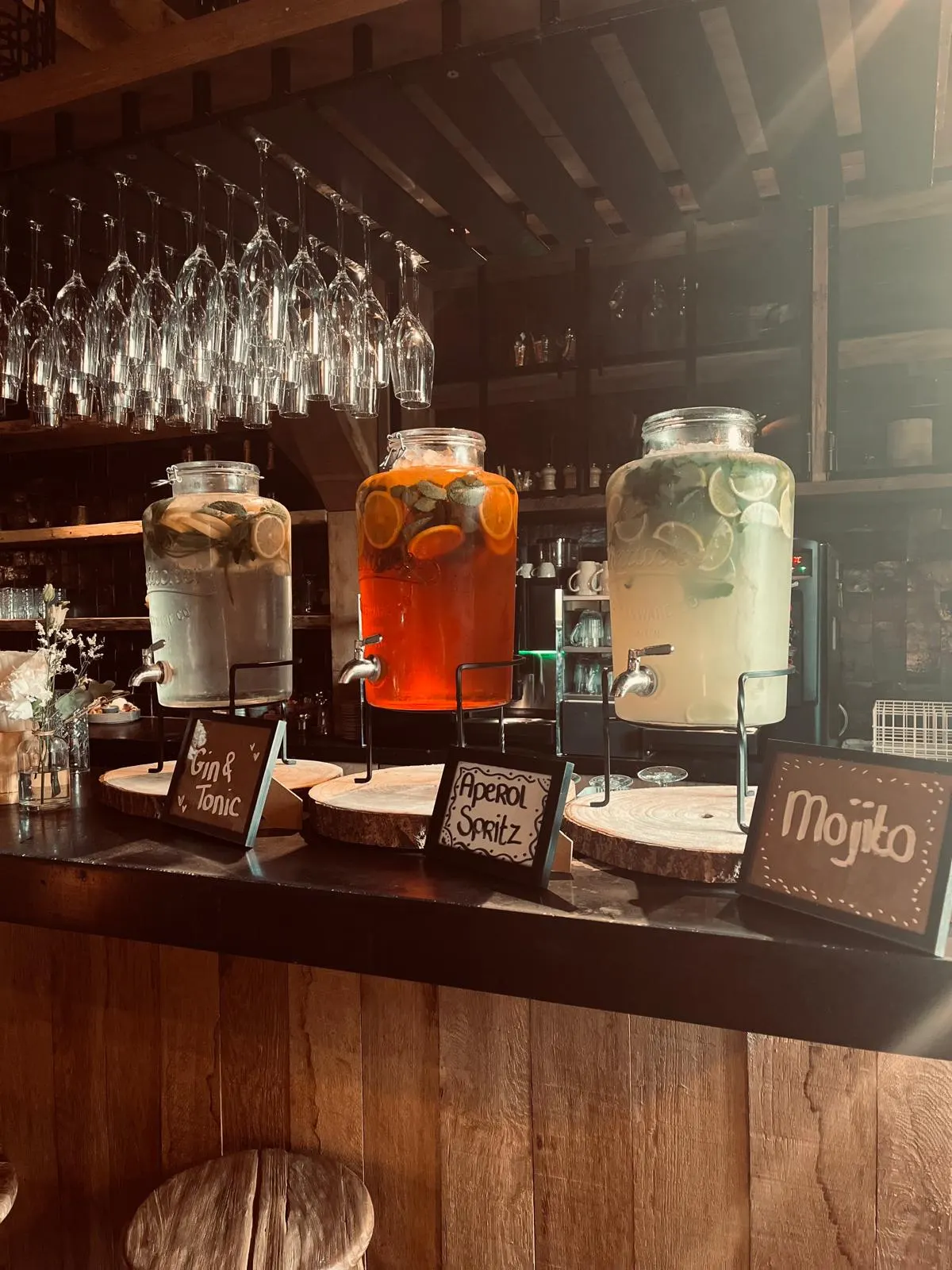 Drie grote glazen drankdispensers op de bar, gevuld met gele, oranje en limoengroene cocktails met schijfjes citrus; ervoor drie houten krijtbordjes met Gin & Tonic, Aperol Spritz en Mojito, en boven de bar rijen wijnglazen ondersteboven.