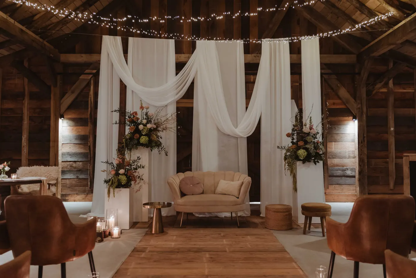 Het avonddecor van de Barn frontaal gezien: witte draperiebanen over de volle hoogte, een crème canapé met een rond kussen ertussen, twee witte sokkels met herfstkleurige bloemstukken, een gouden bijzettafeltje en kaarsen op de grond, met lichtsnoeren onder de kap.