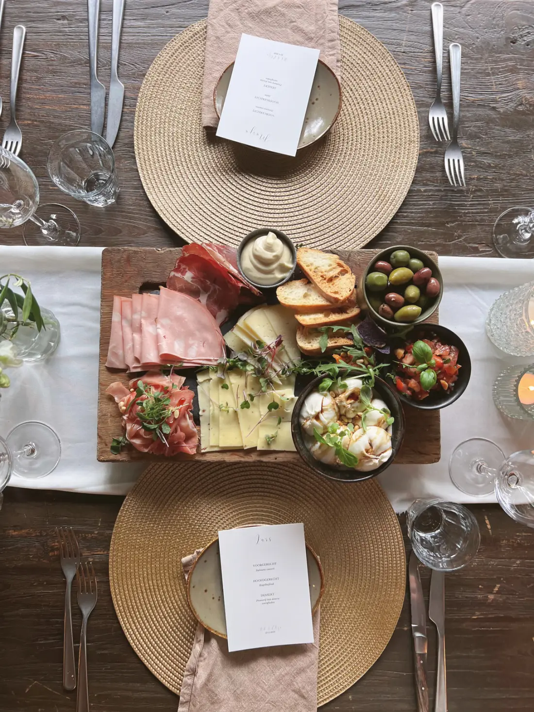 Een deelplank van bovenaf gefotografeerd: dun gesneden ham, kaasblokjes, olijven, tomaatjes, brood en dipjes in schaaltjes, op een witte loper met ronde gevlochten placemats, bestek en een menukaartje ernaast.