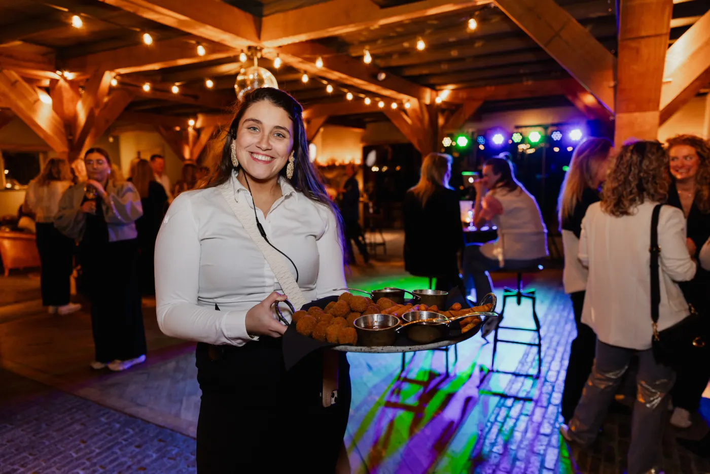 Een lachende serveerster loopt met een dienblad vol glaasjes door de feestzaal, met achter haar dansende gasten in groen en paars podiumlicht onder de houten kap.