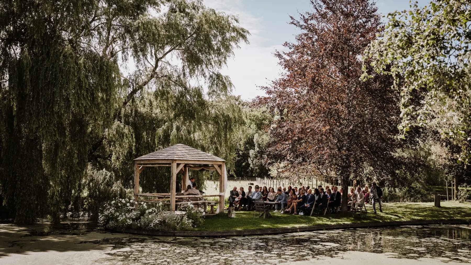 De ceremonie op het eiland, gezien vanaf de overkant van de vijver: het houten prieel met shingledak staat op het gras, rechts ervan twee rijen zittende gasten onder een koperbeuk, links een treurwilg die tot op het water hangt, en op de voorgrond het vijveroppervlak vol kroos en waterlelies.