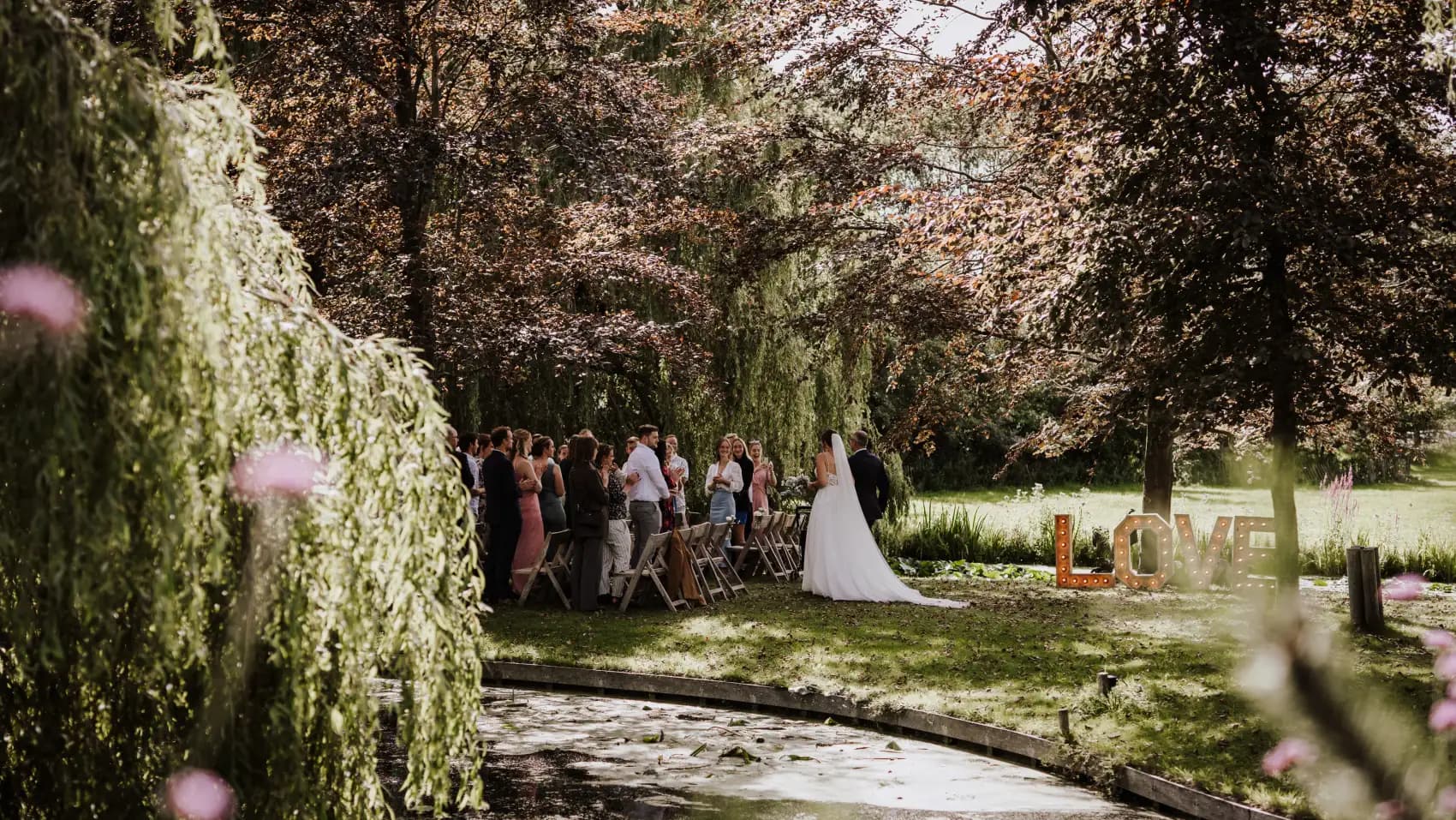 De ceremonie op het eiland gezien door de hangende takken van een treurwilg: gasten staan en zitten langs het water, de bruid loopt met een lange sleep over het gras, en rechts aan de waterkant staan houten LOVE-letters. Op de voorgrond hangen roze bloemen onscherp in beeld.