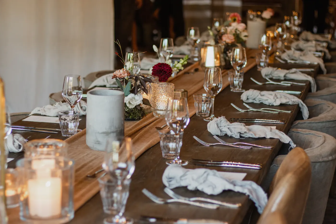 Een lange donkere houten tafel gedekt voor een sit-down diner: wijn- en waterglazen, losjes gevouwen linnen servetten, waxinelichtjes in geribbelde glazen en een klein bloemstuk met een donkerrode dahlia, met bruine leren kuipstoelen langs de tafel.