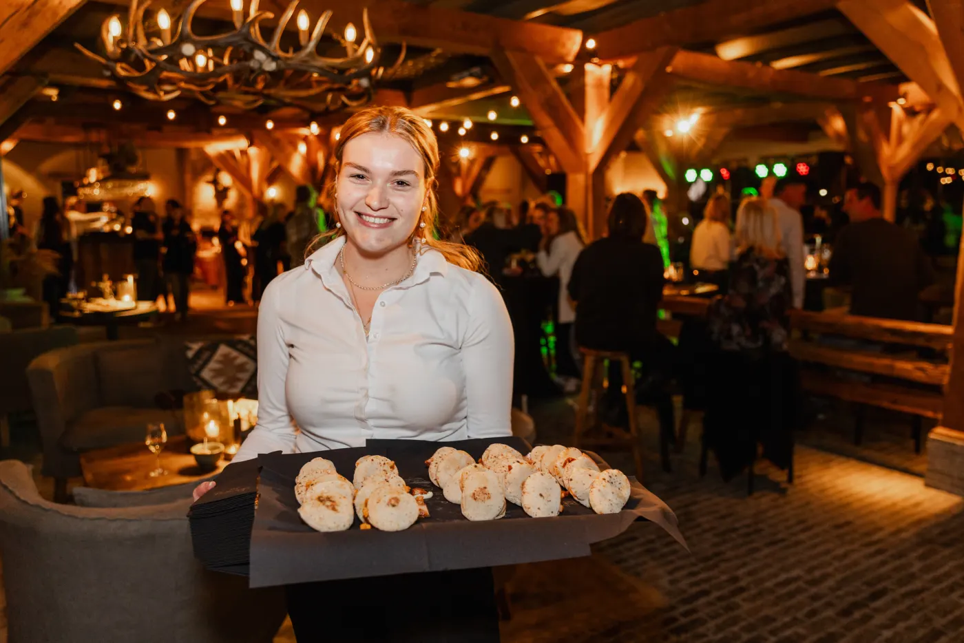 Een lachende serveerster in een witte blouse draagt een zwarte plank met ronde gebakken hapjes door de feestzaal en kijkt recht in de lens; achter haar warme lampjes en de silhouetten van gasten in het donker.