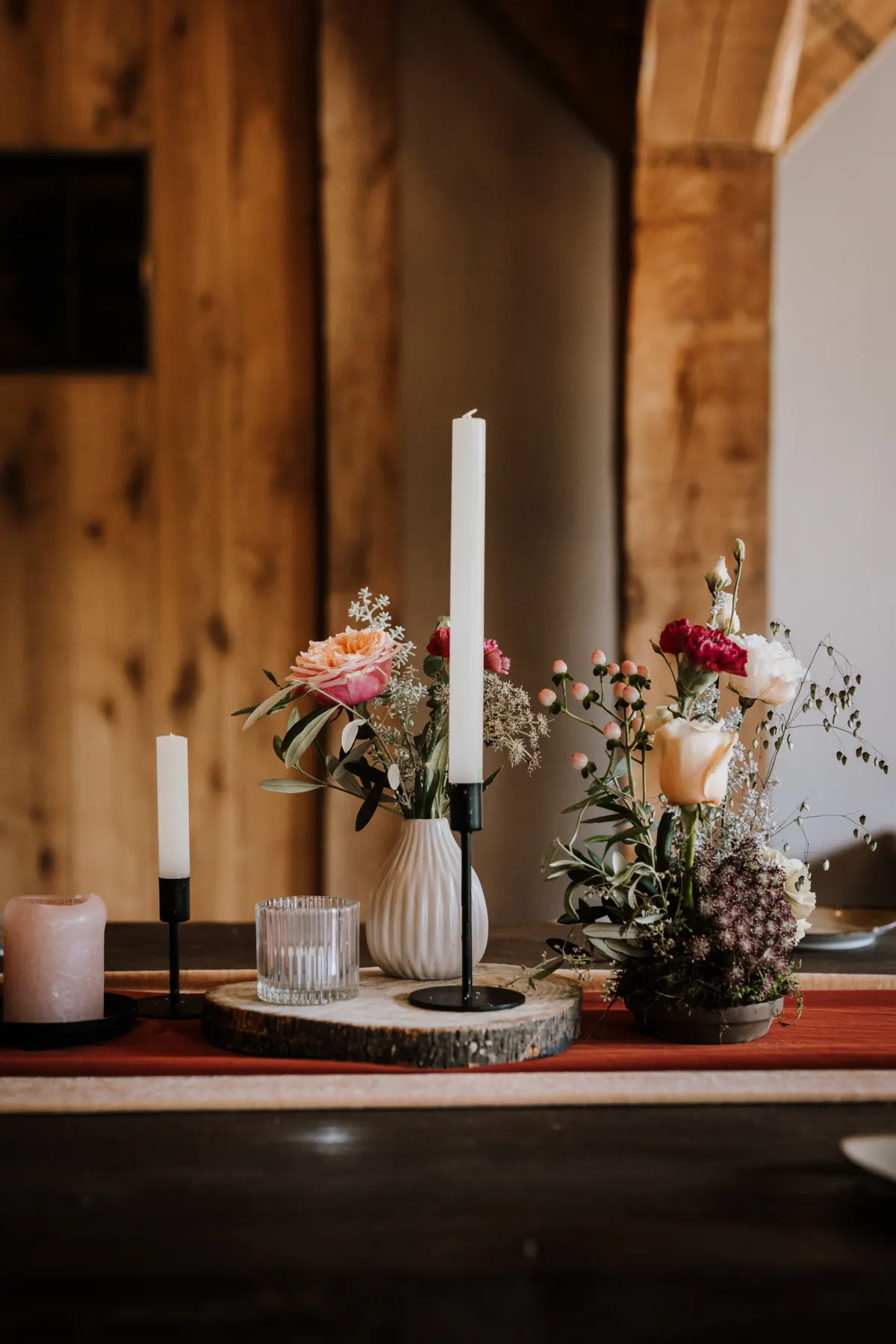 Een tafeldetail tegen een houten wand: twee witte dunne kaarsen in zwarte kandelaars, een geribbeld glas en witte vaasjes met roze, rode en crème tuinbloemen op boomschijven, op een roestrode tafelloper.
