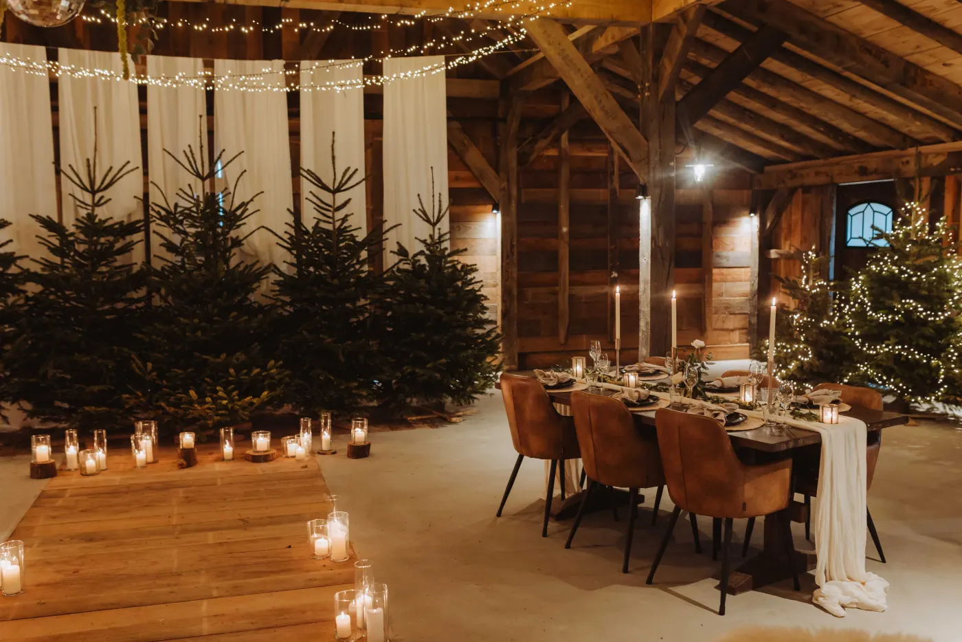 Links een rij echte kerstbomen tegen witte draperiebanen met tientallen kaarsen in glazen op de vloer ervoor, rechts een lange gedekte eettafel met dunne kaarsen in kandelaars, wijnglazen en bruine leren stoelen, en daarnaast een verlichte kerstboom.