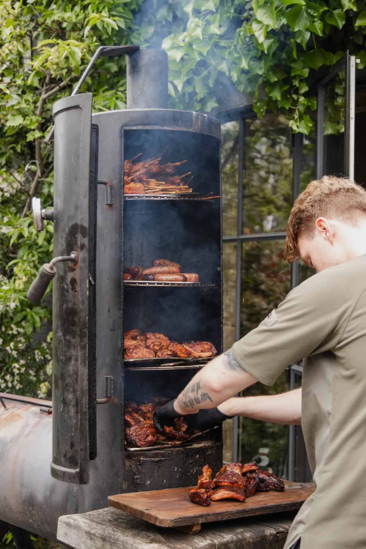 Een jonge kok in een zandkleurig T-shirt trekt buiten een rooster uit een hoge cilindrische rookoven; binnenin liggen spareribs, kippendijen en spiesjes op drie etages, met rook die tussen het groen opstijgt.