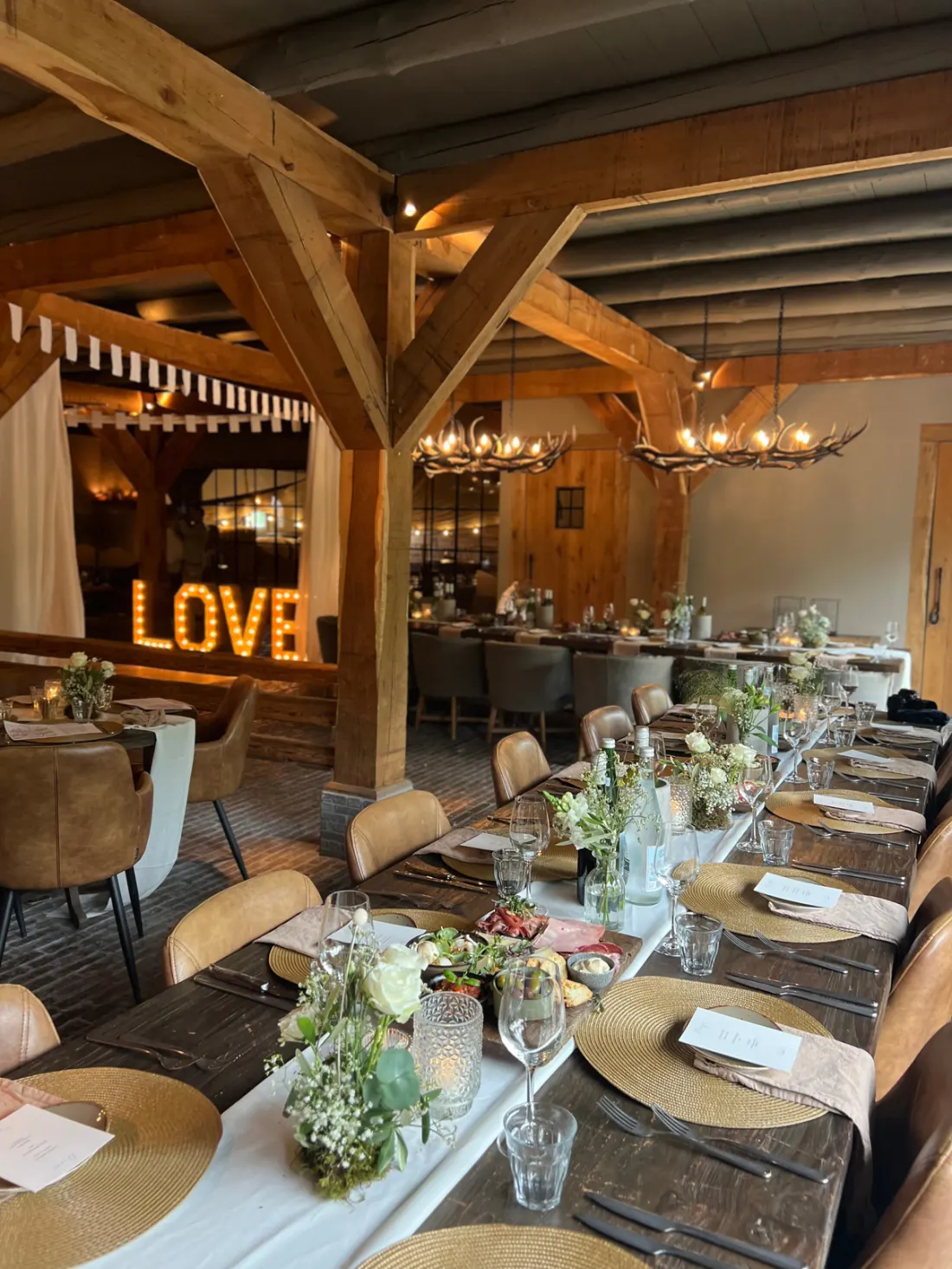 Een lange gedekte tafel met ronde gevlochten placemats, witte bloemstukken en menukaartjes, met op de achtergrond de verlichte LOVE-letters en twee geweikroonluchters onder de houten balken.