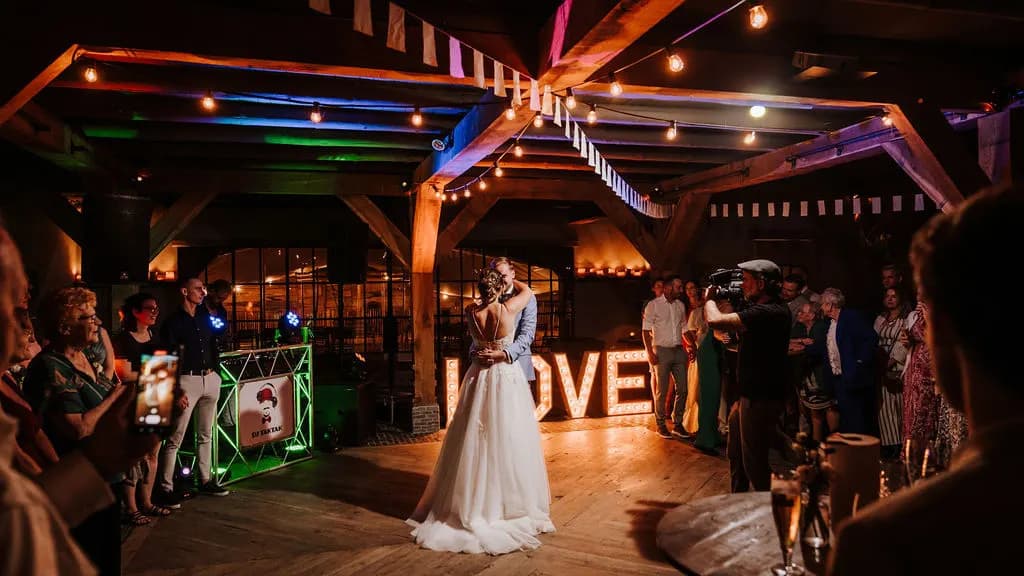 De openingsdans onder festoonverlichting en wimpels: het paar danst midden op de houten vloer voor de verlichte LOVE-letters, omringd door gasten en een videograaf met een camera.
