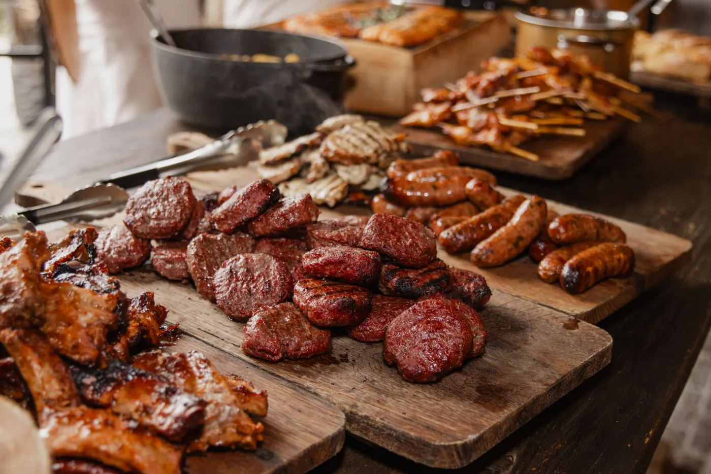 Een houten plank vol gegrild vlees van de barbecue: hamburgers, worsten, spiesjes en gegrilde stukken, met daarachter een zwarte gietijzeren pot en een ovenschaal op de werktafel.