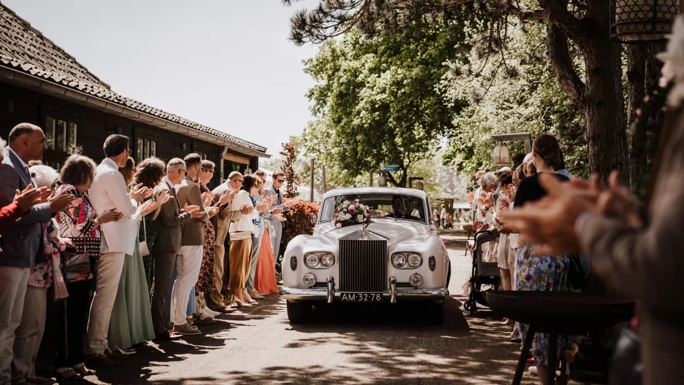 Een witte klassieke Rolls-Royce met bloemversiering op de motorkap staat op de oprijlaan onder hoge bomen; links ernaast een groepje gasten in zomerkleding en een fotograaf die het moment vastlegt.