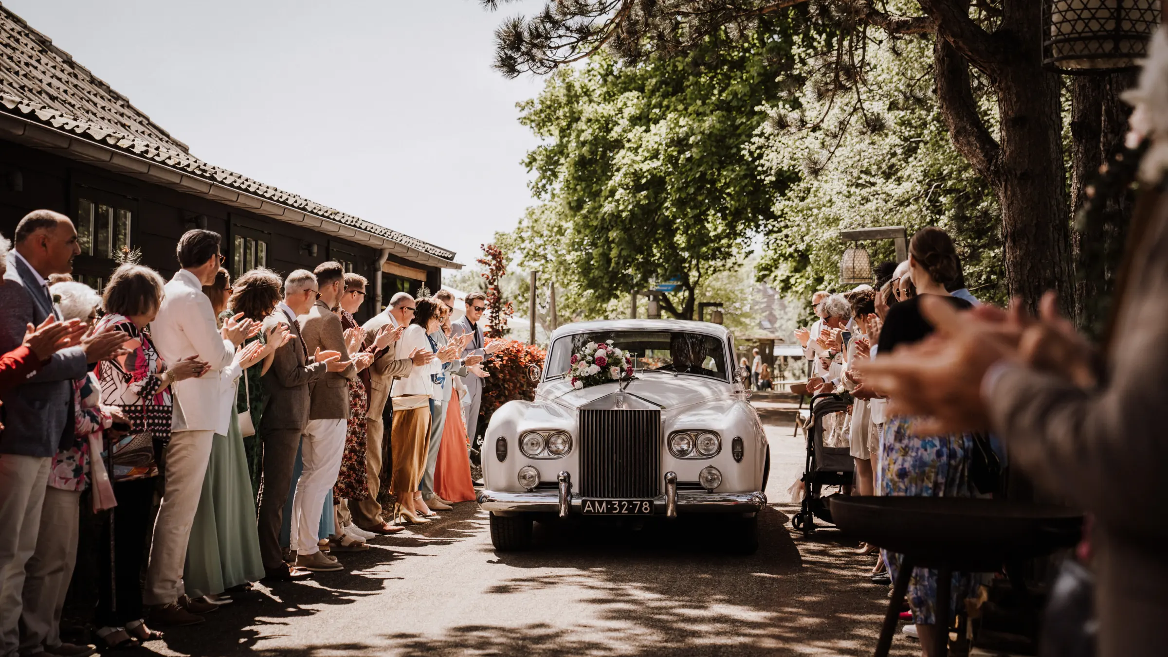 Een witte klassieke Rolls-Royce met bloemversiering op de motorkap staat op de oprijlaan onder hoge bomen; links ernaast een groepje gasten in zomerkleding en een fotograaf die het moment vastlegt.