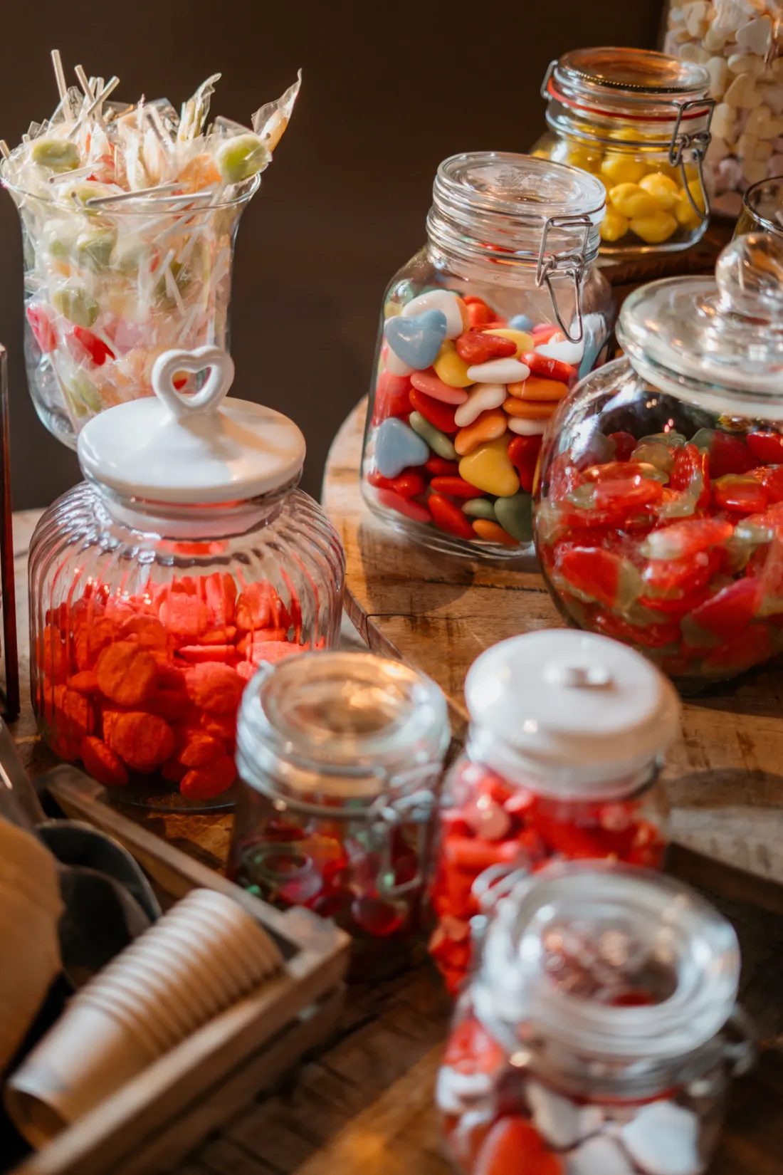 Glazen stopflessen en weckpotten vol snoep op de candy table van dichtbij: rode spekjes, gekleurde zuurtjes en schuimpjes, met papieren zakjes en een schepje op de houten tafel ervoor.