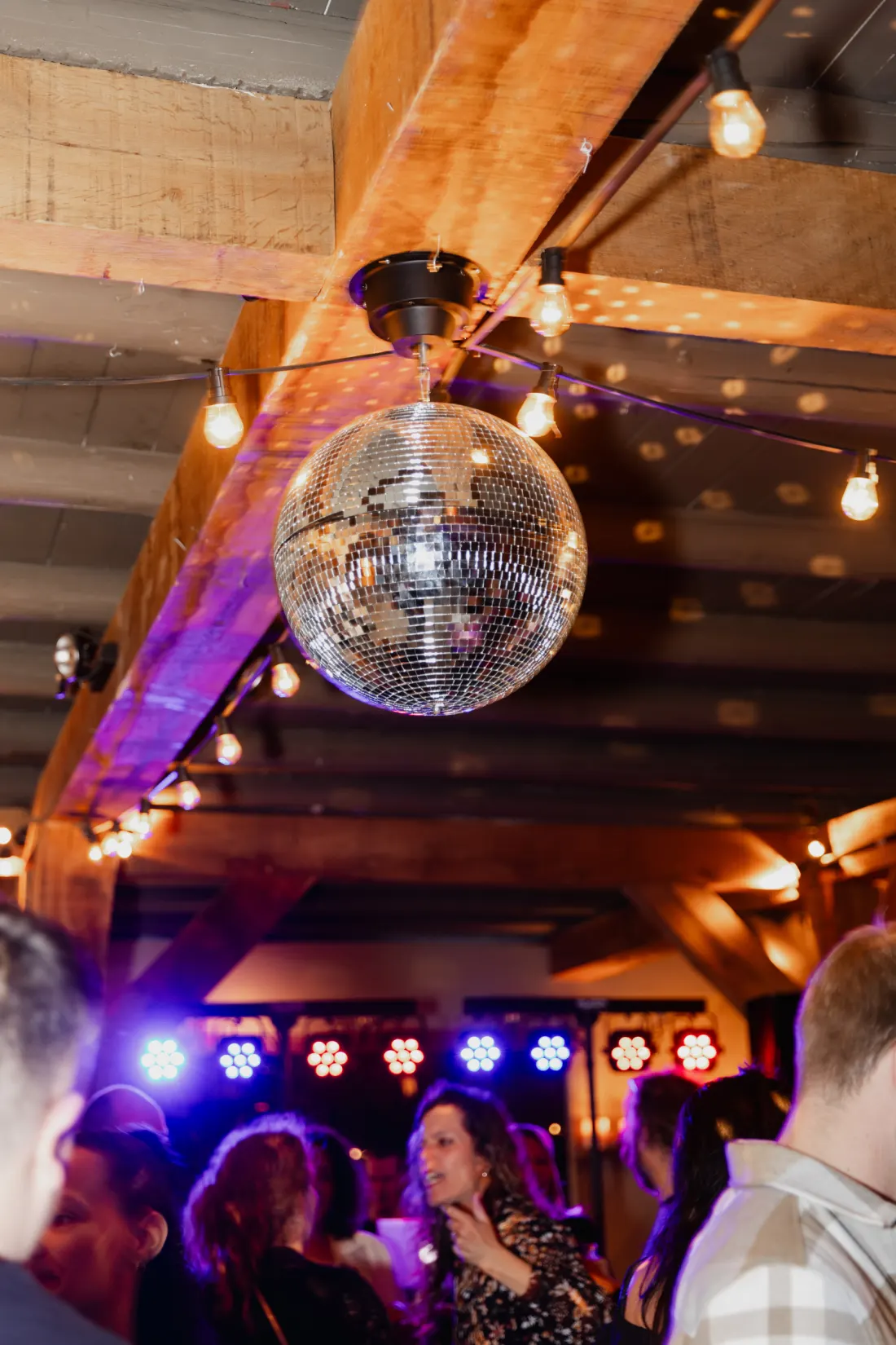 Een spiegelbol hangt aan de houten kap tussen festoonlampjes en werpt lichtstippen over de balken; daaronder een rij gekleurde partylampen en gasten op de dansvloer in het donker.