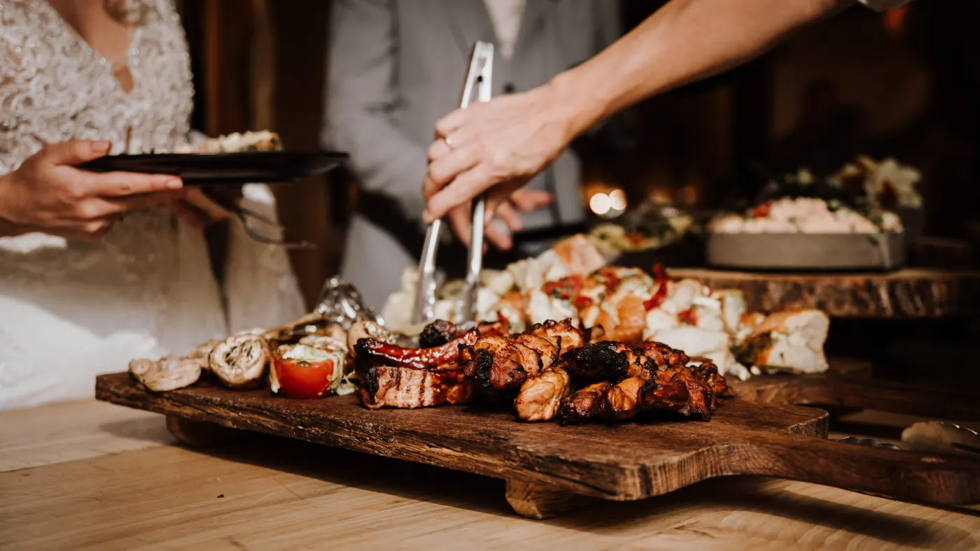 Een hand met een tang pakt gegrilde spiesjes en stukken vlees van een houten plank waarop ook gegrilde champignons en een tomaatje liggen; links ernaast staat een bruid in een kanten jurk met een bord in haar hand.