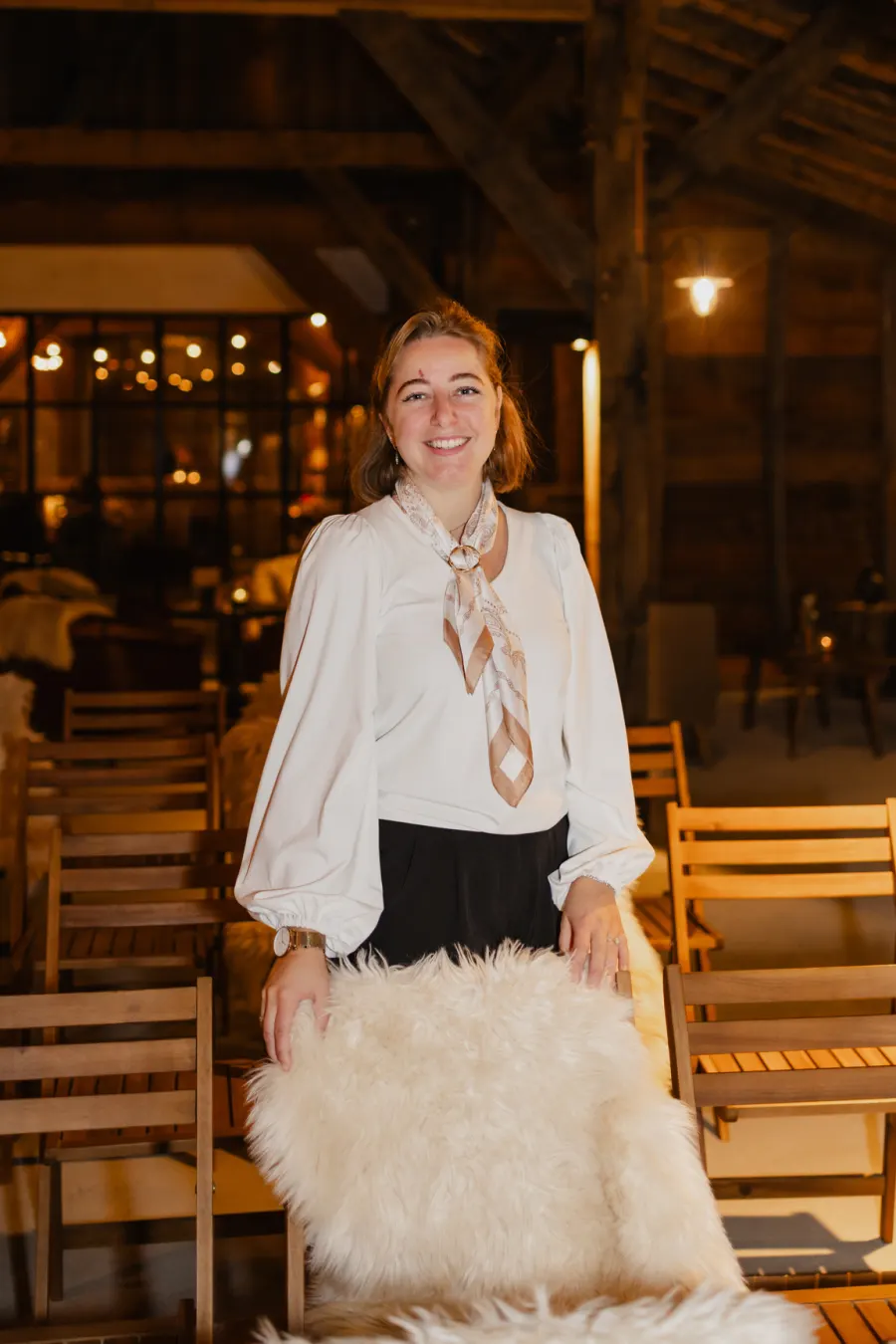 Marlot in een witte blouse met een zijden sjaaltje, leunend op een houten stoel met een schapenvacht erover, met lichtsnoeren onscherp in de schuur achter haar.