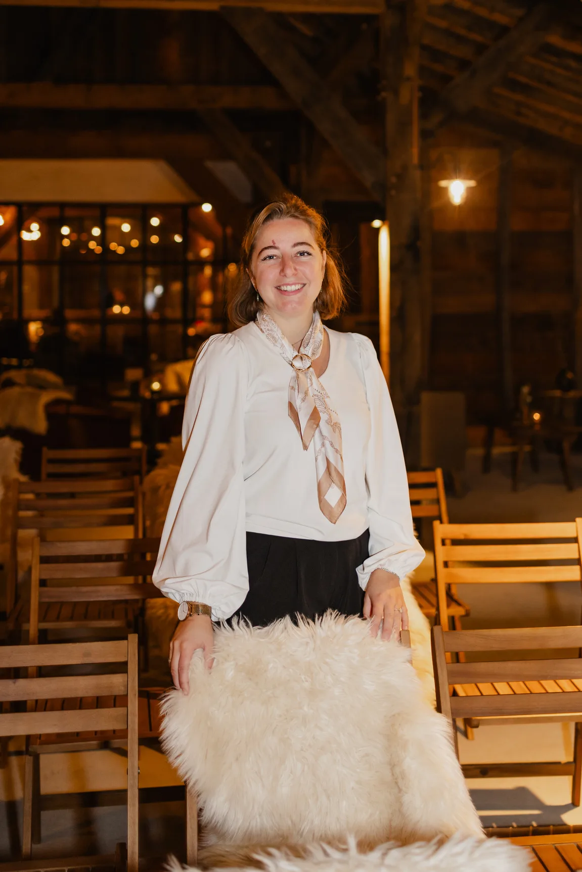 Marlot in een witte blouse met een zijden sjaaltje, leunend op een houten stoel met een schapenvacht erover, met lichtsnoeren onscherp in de schuur achter haar.