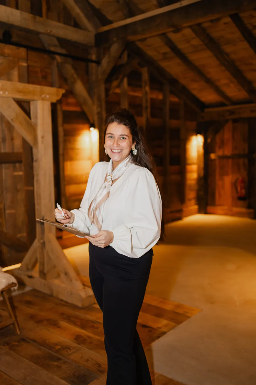 Nikita staat lachend in de schuur met een klembord in haar hand, in een witte blouse en een zwarte broek, tussen de houten balken en het warme kunstlicht.