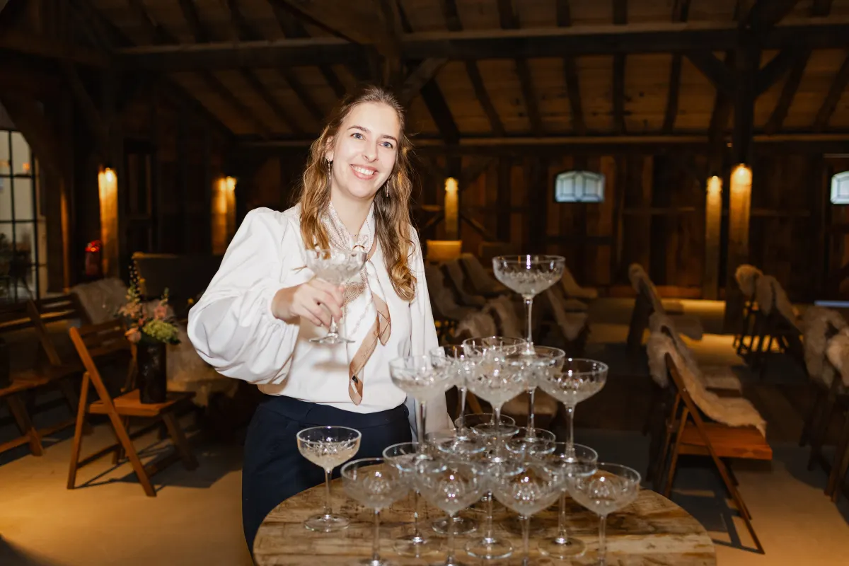 Sanne staat lachend achter een toren van champagnecoupes met een glas in haar hand, in een witte blouse met pofmouwen, met de stoelen en de kap van de schuur achter haar.
