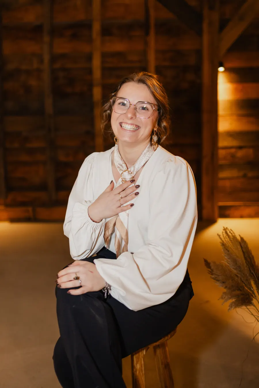 Silvia in een witte blouse met franjes en een bril, haar hand op haar borst en lachend in de lens, met pampasgras rechts achter haar in de schuur.