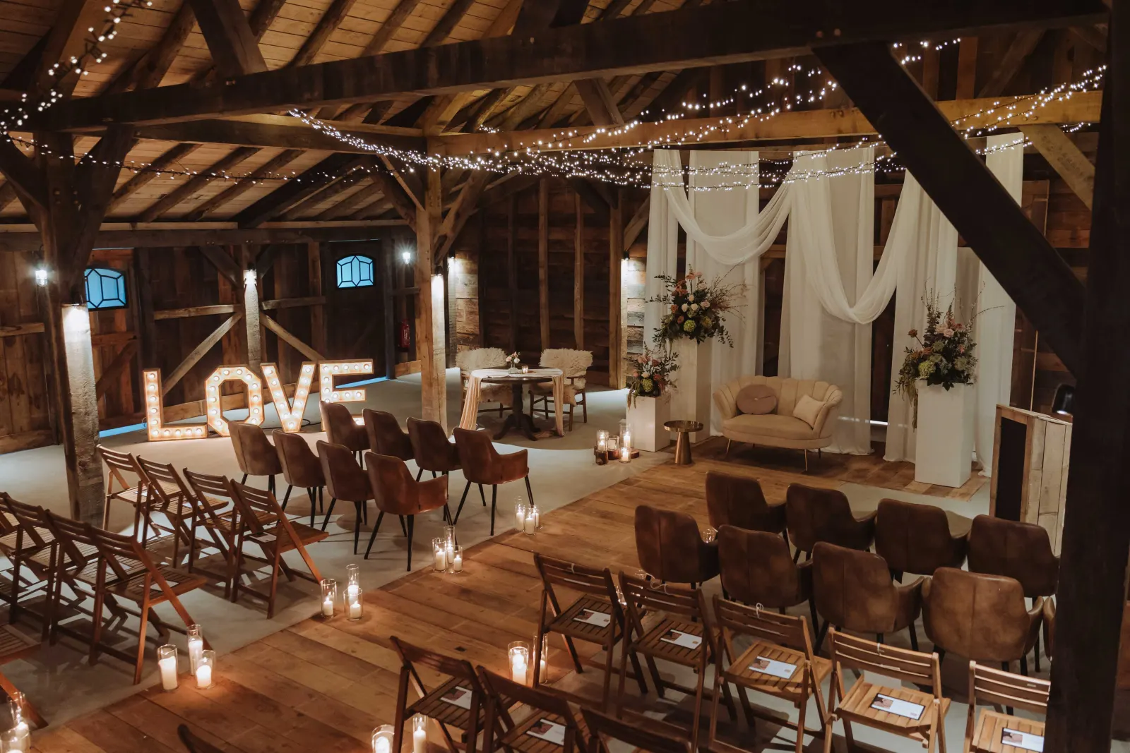 Dezelfde schuur bij avond, met lichtsnoeren kriskras onder de kap: links verlichte marquee-letters die LOVE spellen, daarvoor rijen bruine leren kuipstoelen en houten klapstoelen, en achterin een wit gedrapeerd decor met herfstkleurige bloemstukken op witte sokkels en een crème canapé.