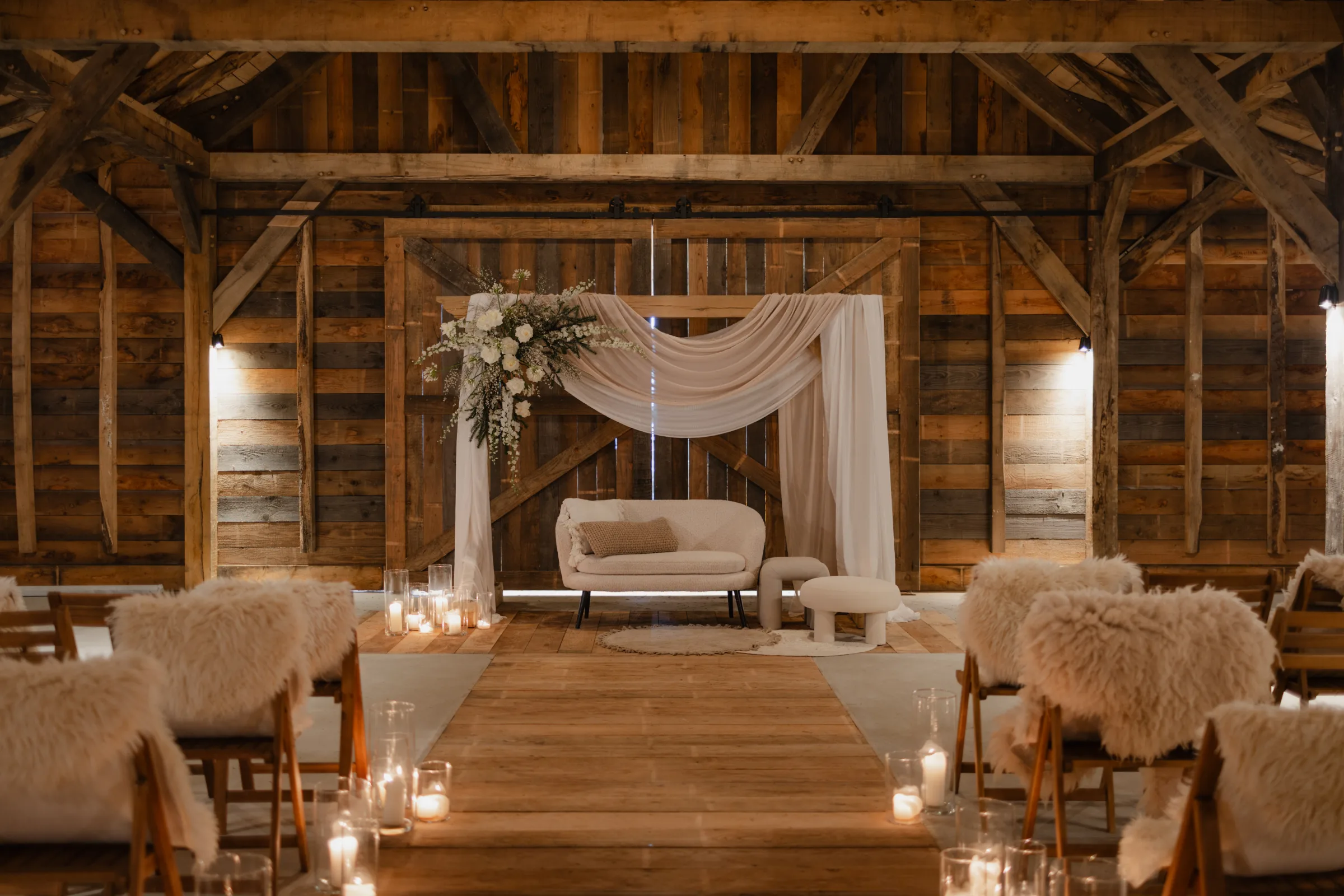 De Barn ingericht voor een ceremonie: donker verweerd hout aan wanden en spanten, achterin een witte gedrapeerde boog met een wit bloemstuk ernaast en een crème tweezitsbank op een rond jute kleed. Een houten loper vormt het middenpad, met kaarsen in glazen op de vloer en klapstoelen met schapenvachten aan weerszijden.