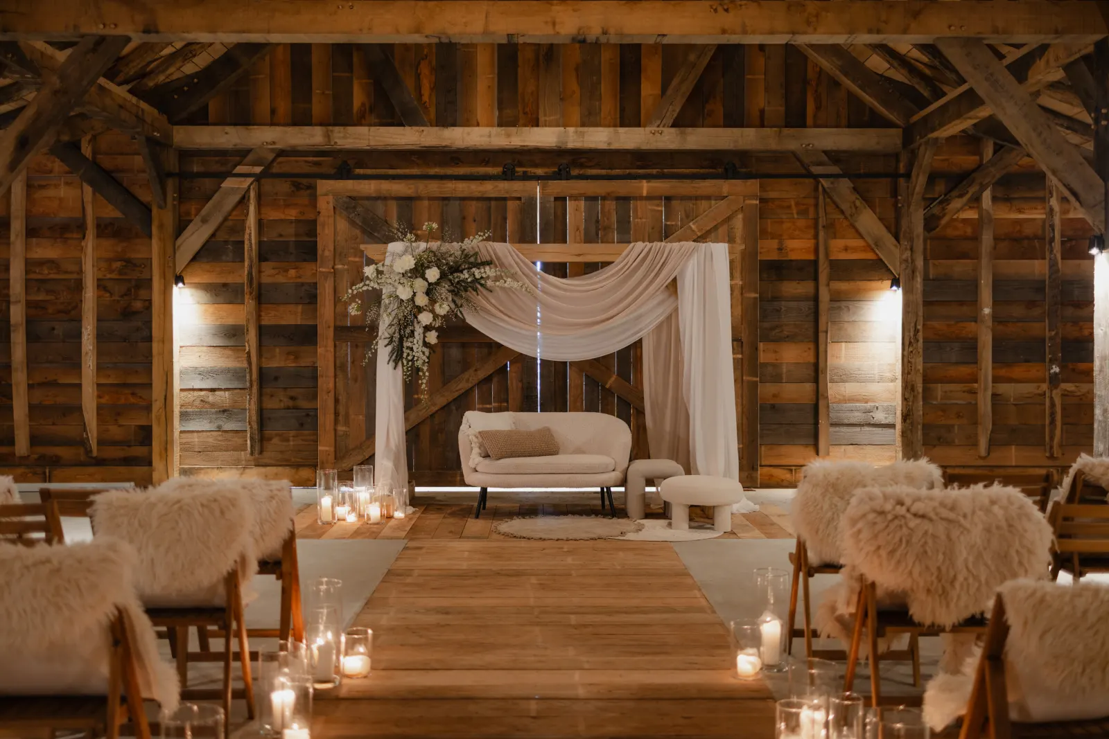 De Barn ingericht voor een ceremonie: donker verweerd hout aan wanden en spanten, achterin een witte gedrapeerde boog met een wit bloemstuk ernaast en een crème tweezitsbank op een rond jute kleed. Een houten loper vormt het middenpad, met kaarsen in glazen op de vloer en klapstoelen met schapenvachten aan weerszijden.
