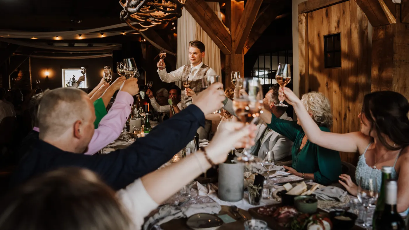 Een toost aan de lange tafel: een tiental gasten heft de wijn- en waterglazen met gestrekte armen boven de tafel, en achterin staat een jonge man lachend rechtop met zijn glas omhoog onder een geweikroonluchter.
