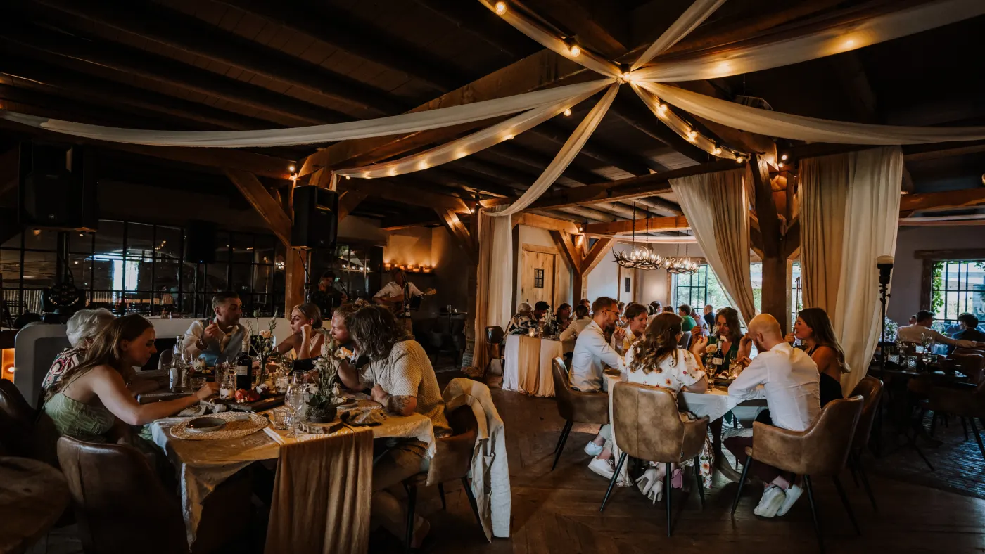 De volle zaal aan tafel: gasten zitten aan ronde en lange tafels onder crème stofbanen die stervormig over de houten kap gespannen zijn, met lichtsnoeren en geweikroonluchters erboven.