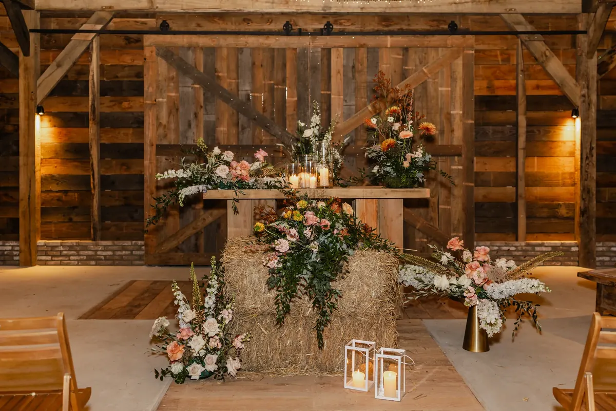 Een hooibaal tegen de houten wand van de binnenstal, overgroeid met een bloemstuk van roze en oranje rozen, wit fluitenkruid en groen. Op de balk erboven staan brandende kaarsen tussen twee bloemstukken; op de vloer ervoor twee lantaarns met kaarsen en losse bossen witte en roze bloemen.