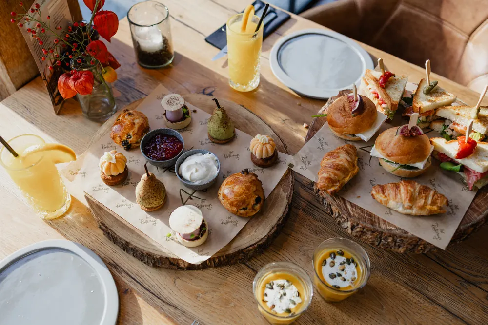 Twee boomschijven naast elkaar op een lange eiken tafel. Op de linker het zoet: een rozijnenscone, taartjes met een toef room in de vorm van een pompoentje, een glanzend groen dessert als een peer, en kommetjes met rode jam en slagroom. Op de rechter het hartige: croissants, zachte broodjes met een halve vijg op een prikker, en geroosterde sandwiches met tomaat en sla. Vooraan twee glaasjes oranje soep met room en pitten en twee glazen jus d’orange met citroen, achteraan een vaasje met oranje lampionnetjes en een kaartje, en aan weerszijden lege grijze borden.