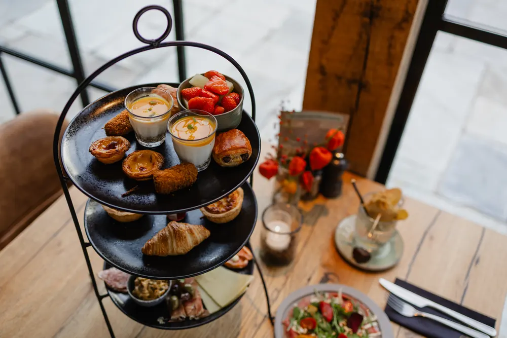 Een zwarte etagère van drie schalen bij een stalen raampui die uitkijkt op de bestrating buiten. Bovenop het zoet en het warme: custardtaartjes, een krokante kroket, twee glaasjes met pannacotta en een oranje mousse, een bakje aardbeien met meloen, en een chocoladebroodje. Op de middelste schaal een croissant en kleine taartjes, op de onderste charcuterie, kaas en olijven. Ernaast een waxinelichtje, een vaasje met oranje lampionnetjes, een staand menukaartje, een cocktail met een gedroogde schijf fruit, en een bord met salade naast bestek op een donkerblauw servet.