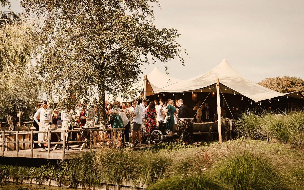 Gasten met glazen in de hand op de houten vlonder aan het water, voor twee witte stretchtenten met puntige masten en festoenverlichting langs de rand. Links een berk en een treurwilg over de vlonder, op de voorgrond de oevergrassen langs de vijver.