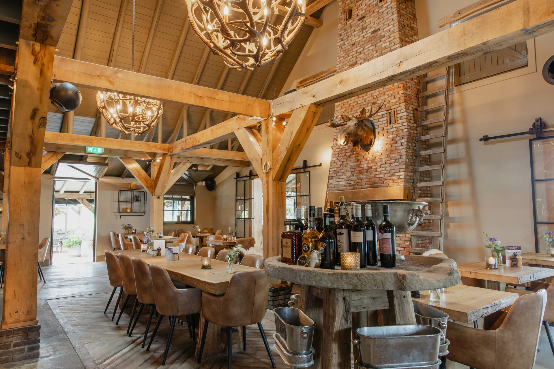 De restaurantzaal van onder de kap: twee kroonluchters van hertengeweien hangen aan zware eiken spanten. In het midden een ronde tafel van een boomstamschijf met wijnflessen en een zilveren koelvat erop en zinken koelemmers eronder; links een lange eiken tafel met cognac leren stoelen en een schuifpui die open staat naar het overdekte terras. Rechts een gemetselde schoorsteen met een elandkop en een houten ladder ertegenaan.