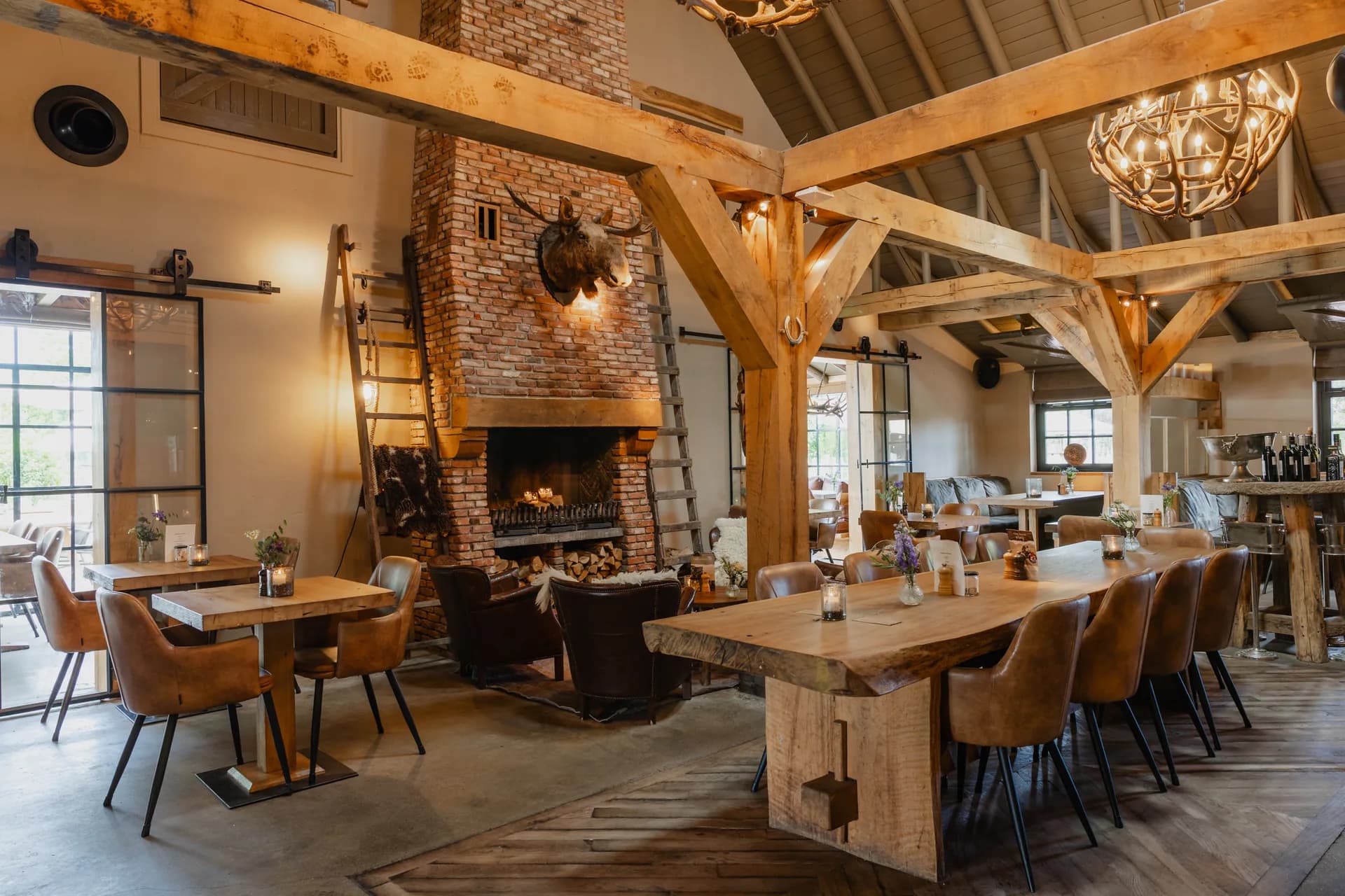 De restaurantzaal met de open haard: in een gemetselde schouw met een zware eiken balk brandt een houtvuur, met een elandkop op de schoorsteen erboven en een houten ladder ertegenaan. Ervoor twee donkerbruine leren fauteuils op een kleed; links kleine eiken tafels met cognac leren stoelen bij een stalen pui, rechts een lange eiken tafel met kaarslicht en veldbloemen onder de eiken spanten en een kroonluchter van hertengeweien.