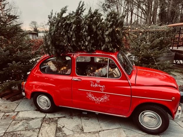 Ein roter Fiat 600 mit einem Weihnachtsbaum auf dem Dach und „Het Ruiterhuys“ in weißen Buchstaben auf der Tür, geparkt auf dem Natursteinboden zwischen Weihnachtsbäumen mit Lichtern.
