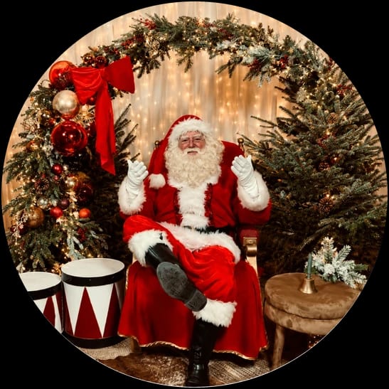 De kerstman in een rode fauteuil, lachend met zijn witte handschoenen omhoog, onder een boog van dennengroen met rode kerstballen en een grote rode strik; naast hem kerstbomen met lichtjes en twee trommels in rood en wit.